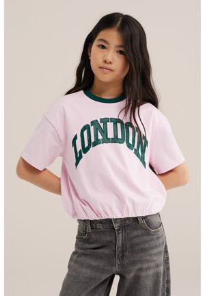 Ung pige med langt mørkt hår iført en lyserød T-shirt med "LONDON" i grønne bogstaver og mørkegrå jeans mod en ensfarvet baggrund.