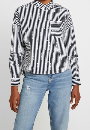 Schwarz-weiß gestreiftes Hemd mit Knöpfen und vertikalen Streifen, bedruckt mit dem Schriftzug "Tommy Jeans", kombiniert mit hellblauen Jeans, die eine kleine Used-Detailierung aufweisen.