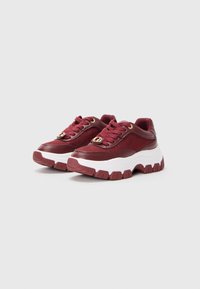 Une paire de baskets chunky bordeaux avec de grosses semelles blanches, des côtés en tissu texturé et des accents dorés sur les lacets, sur un fond gris.