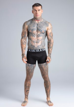 Raumeningas vyras su plačiais viso kūno tatuiruotėmis, įskaitant didelį veidą ant krūtinės, dėvi juodus bokserius, stovi prieš paprastą pilką foną.