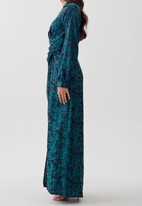 Lång, tealblå blommig klänning med långa ärmar, en sidofalsk och ett knutet midjedesign, tillverkad av mjukt tyg.