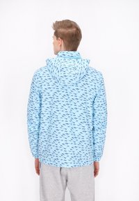 Hellblaue Jacke mit einem sich wiederholenden schwarzen Muster kleiner Pferde, ausgestattet mit einer Kapuze und elastischen Bündchen. Glatte Textur, leichtes Design.