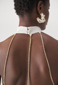 Dossier d'une robe sans manches blanche avec un col haut, agrémentée de deux chaînes en strass dorés qui tombent et d'une fermeture à bouton au niveau du col.