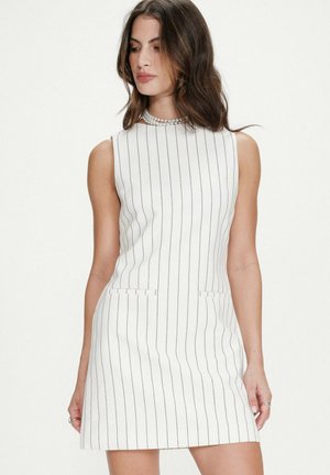 Femme portant une robe blanche sans manches avec de fines rayures noires verticales et un collier de perles, se tenant devant un fond uni.