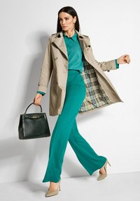 Vrouw loopt in een teal blouse en broek, beige trenchcoat met geruite voering, beige hakken, en draagt een zwart gestructureerde handtas.