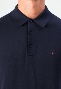 Námornícky modré polo tričko vyrobené z textúrovanej tkaniny. Má plochý golier, zapínanie na tri gombíky a malú logo v červenej, bielej a modrej farbe na hrudi.