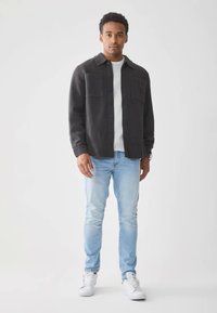 Hombre vestido con una chaqueta oscura de botones, sudadera gris clara, jeans azul claro y zapatillas blancas, de pie contra un fondo blanco liso.