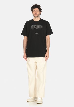T-shirt in cotone nero con il logo "MOSCHINO" e "Milano" in bianco, abbinata a pantaloni a gamba larga crema e sneakers bianche.