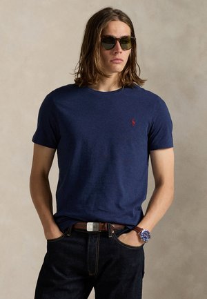 Giovane uomo con capelli lunghi che indossa occhiali da sole scuri, una t-shirt blu navy, jeans scuri, una cintura marrone e un orologio con quadrante blu.