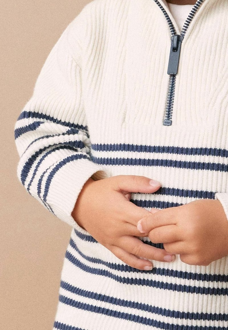 Enfant portant un pull côtelé blanc avec des rayures bleu marine et une demi-fermeture éclair, tenant ses mains jointes à la taille.