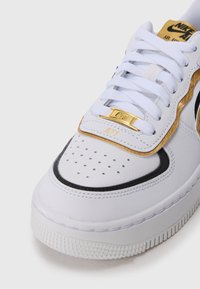 Białe sneakersy Nike Air Force 1 z złotymi akcentami, białymi sznurówkami, perforowanym noskiem i czarnymi wykończeniami na białym tle.