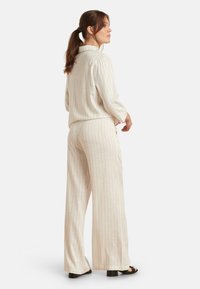 Licht beige twee-delige outfit met verticale strepen; kraagtop met lange mouwen en losse, wijde broekspijpen; textiel met structuur; haar van gemiddelde lengte.