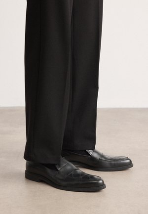 Schwarze Leder-Loafers mit glatter Oberfläche und geprägtem Muster. Sie zeichnen sich durch ein flaches Profil und dezente Nahtakzente aus. Kombiniert mit schwarzen Hosen.