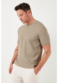 Khaki színű, bordázott, rövid ujjú póló kerek nyakkal, texturált anyaggal és laza fazonnal, világos színű nadrággal párosítva.