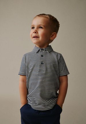 Ung dreng med kort hår iført en navy- og hvidstribet poloshirt og navy bukser, ser op med hænderne i lommerne mod en ensfarvet baggrund.