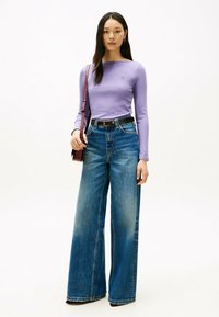 Hellblau gestreiftes Langarmoberteil in Lila, kombiniert mit hoch taillierten, weit geschnittenen blauen Jeans. Das Model hält eine kleine dunkle Tasche und zeigt einen schlichten Gürtel.