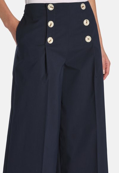Tengerészkék, magas derekú, széles szárú nadrág hat nagy fehér gombbal és oldalzsebekkel, fehér betűrt felsővel viselve.