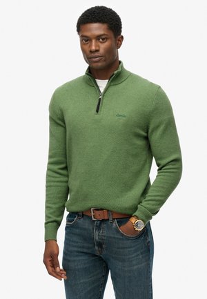 Jersey verde de punto con cuello de media cremallera, tejido texturizado y logo bordado. Combinado con jeans de mezclilla azules y un cinturón marrón.