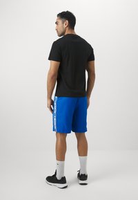 Μαύρη κοντομάνικη μπλούζα, μπλε σορτς με λευκή πλευρική ρίγα, και μαύρα αθλητικά παπούτσια με γκρι κάλτσες, από πίσω σε λευκό φόντο.