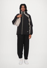 Veste noire avec des rayures blanches, fermeture éclair, col montant, associée à un pantalon noir ample et des baskets blanches. Le mannequin porte un sac beige.