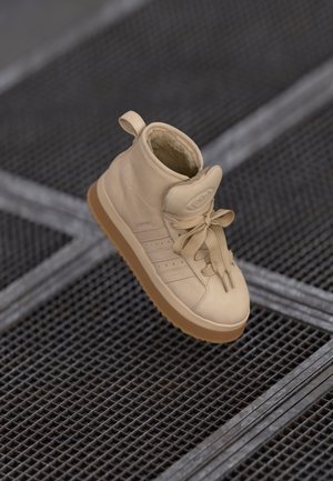 Beige højtop sneaker med snørebånd og varm inderfor, hvilende på en metalrist overflade.