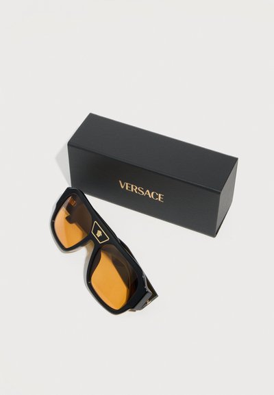 Μαύρα και χρυσά γυαλιά ηλίου Versace με κεχριμπαρένιους φακούς δίπλα σε μαύρο κουτί Versace με χρυσά γράμματα πάνω σε λευκή επιφάνεια.