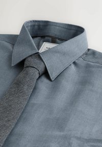 Helles, strukturiertes, himmelblaues Hemd mit umgeschlagenem Kragen, kombiniert mit einer grau gemusterten Krawatte. Etikett im Kragen sichtbar.