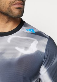 Camiseta deportiva en gris con un diseño estampado. Presenta un cuello negro y un logo azul en el hombro. La tela tiene una textura acanalada.