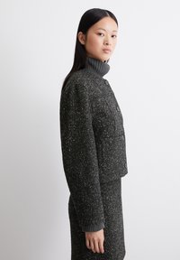 Grau meliertes Jacket mit hohem Kragen, Knopfleiste, zwei Brusttaschen und gerippten Bündchen. Strukturierte Oberfläche mit einem dezenten Muster.