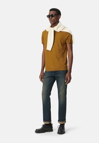 T-shirt marrone a maniche corte, maglione crema chiaro drappeggiato, jeans di denim blu, scarpe nere e occhiali da sole. Il modello è in piedi contro uno sfondo bianco.