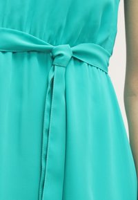 Robe turquoise avec une ceinture en tissu nouée à la taille, montrant des détails froncés et une texture lisse sur un matériau léger.
