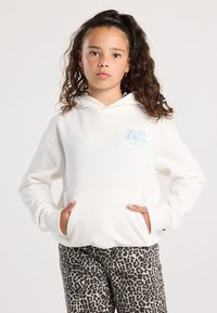 Ragazza con capelli ricci che indossa un hoodie bianco con il testo "The City of Dreams New York" e pantaloni con stampa leopardata, mani nella tasca del hoodie.