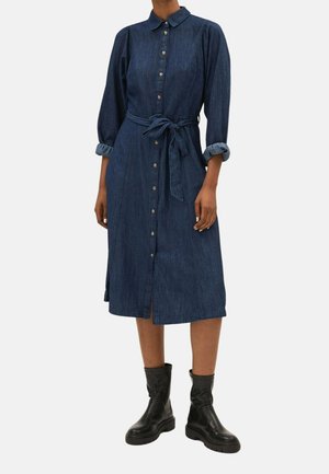 Mujer con vestido midi de mezclilla azul oscuro con botones, mangas largas, cinturón en la cintura y botines negros, de pie frente a un fondo liso.