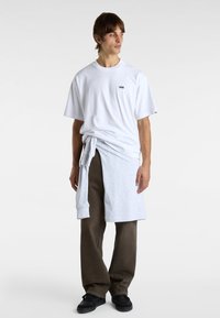 T-shirt en coton blanc avec un petit logo noir, associé à un pantalon de survêtement gris noué à la taille. Le modèle porte des baskets noires. Fond neutre.