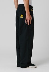 Pantalon noir en tissu résistant, avec une coupe ample, des poches latérales et un patch graphique jaune. Design simple avec une quincaillerie minimale.