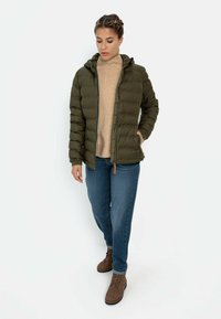 Olive green padded jacket with a hood, front zip, and side pockets, paired with a beige sweater, blue jeans, and brown boots.