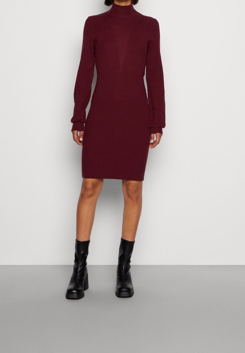 Robe en maille côtelée bordeaux avec col montant, silhouette ajustée et manches longues ; associée à des bottines noires à talons.