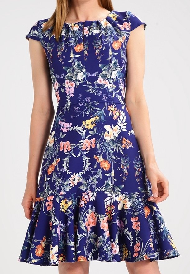 Femme portant une robe bleue à fleurs avec des manches courtes et un ourlet évasé à volants, debout devant un fond uni.
