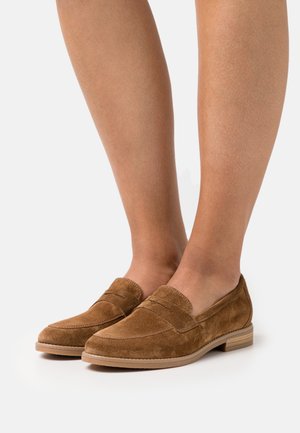 Mocassins en daim marron avec un petit talon, arborant des accents cousus et un design de mocassin. La texture est douce, améliorant confort et style.