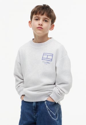 Dreng iført lysegrå sweatshirt med blåt logo og blå jeans med hænderne i lommerne, mod en ensfarvet hvid baggrund.