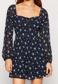 Mörkblå blommig klänning i lätt, genomskinligt tyg. Har fyrkantig halsringning, långa puffärmar, dragsko-detalj och volangfåll.