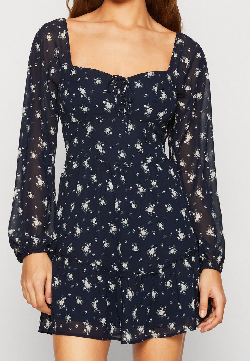 Mörkblå blommig klänning i lätt, genomskinligt tyg. Har fyrkantig halsringning, långa puffärmar, dragsko-detalj och volangfåll.