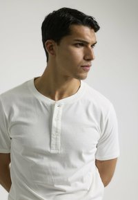 Camisa henley blanca de algodón de manga corta con una tapeta de tres botones y un cuello redondeado. Textura suave, diseño minimalista.