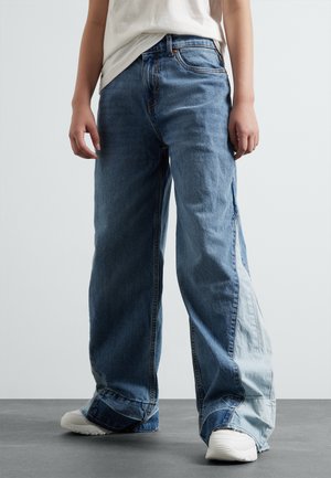 Jean en denim bleu à jambes larges avec un panneau contrasté de teintes plus claires, des ourlets effilochés et un design classique à cinq poches.