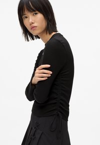 Czarny longsleeve z długim rękawem wykonany z prążkowanej tkaniny, z marszczonymi bokami i detalami w postaci wiązań. Posiada okrągły dekolt i dopasowany krój.