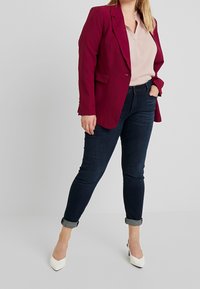 Blazer color marrone, blusa rosa chiaro, jeans skinny blu scuro e tacchi bianchi a punta. Il blazer ha un singolo bottone e maniche leggermente arrotolate.