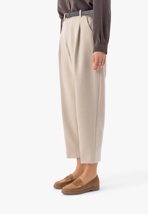 Vrouw die beige cropped broek draagt, een bruin blouse met lange mouwen ingestopt, zwarte riem en bruine suède loafers, staand in zijaanzicht.