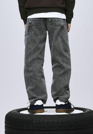 Pantalones cargo de mezclilla gris con un corte holgado, que presentan bolsillos traseros y costuras visibles, combinados con zapatillas azul marino sobre un neumático.