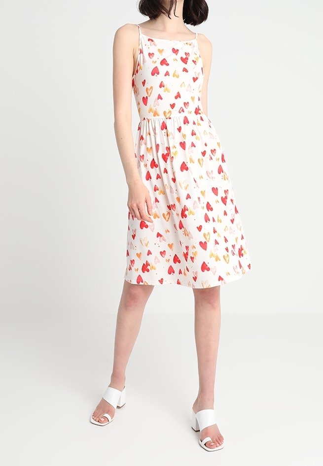 Robe blanche sans manches avec une taille froncée, présentant un motif de cœurs colorés en rouge et doré. Portée avec des sandales à talons blanches.