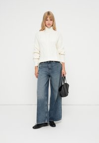 Pulover din cașmir groasă cu guler tip turtleneck, cu model împletit, combinat cu blugi albaștri cu croială largă și o geantă mică din piele neagră.